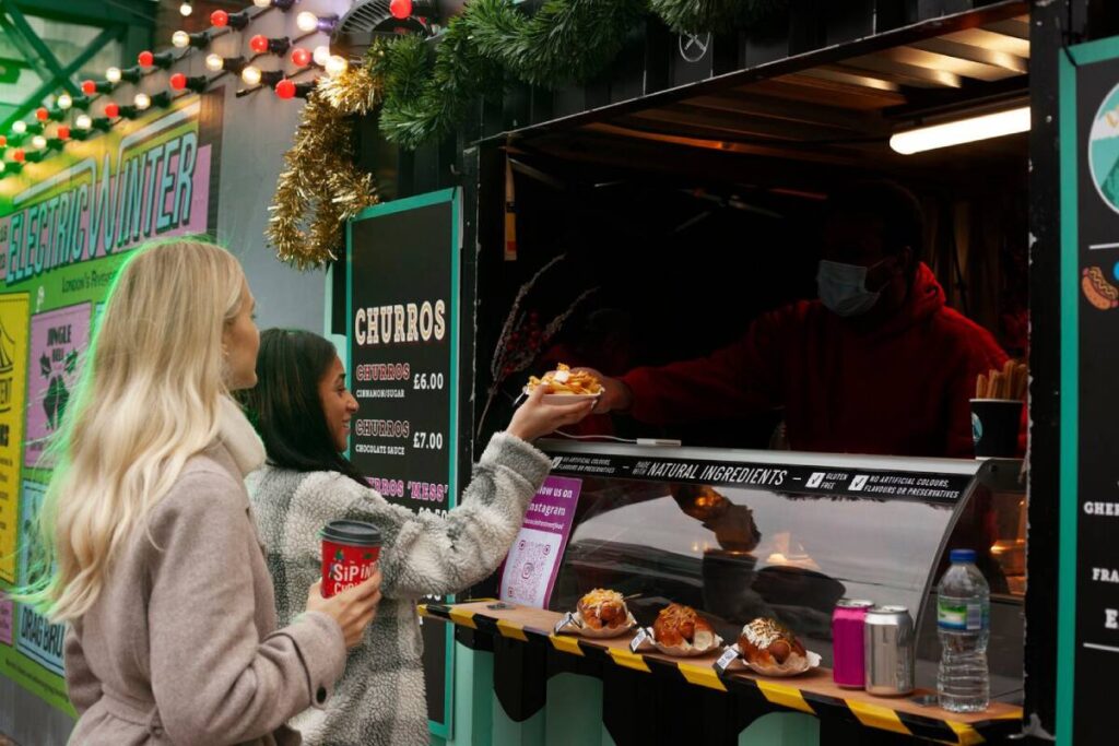 Two women buying food from a street vendor at a festive outdoor market.