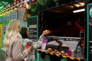 Two women buying food from a street vendor at a festive outdoor market.