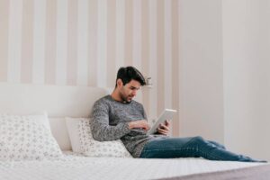 Man sitting on a bed reading a tablet in a striped-wall room.