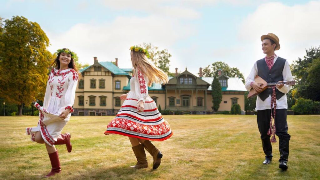 Two women in traditional embroidered dresses dancing joyfully while a man plays a mandolin, in a sunny park in front of a historic house.