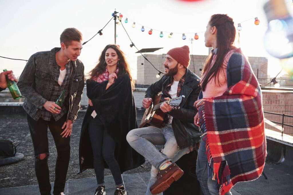 Four young adults enjoying a rooftop gathering with music, drinks, and festive lights at sunset.