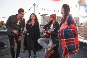 Four young adults enjoying a rooftop gathering with music, drinks, and festive lights at sunset.