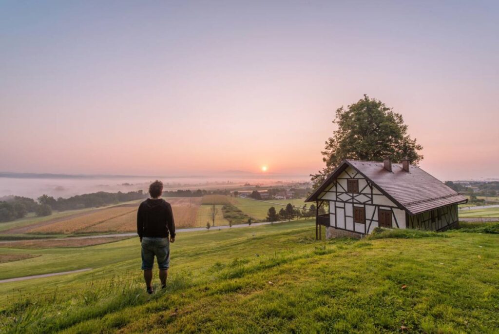 A man standing in a grassy field watching a sunrise near a traditional half-timbered house.