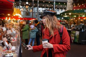 Woman in red coat and black cap using phone and holding coffee cup at a busy market.