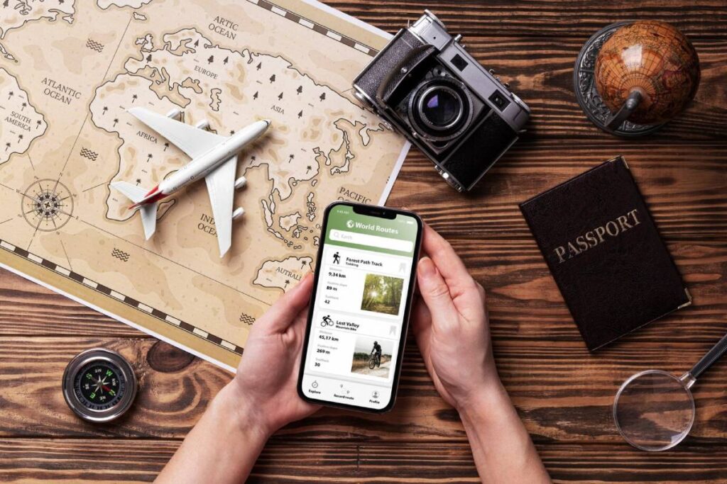 Hands holding a smartphone over a travel map with a passport, plane, camera, and compass on a wooden table.