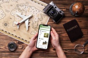 Hands holding a smartphone over a travel map with a passport, plane, camera, and compass on a wooden table.