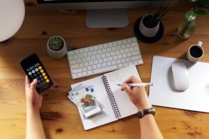 Person managing budget with calculator and writing in notebook on a wooden desk, surrounded by computer, plants, and coffee.