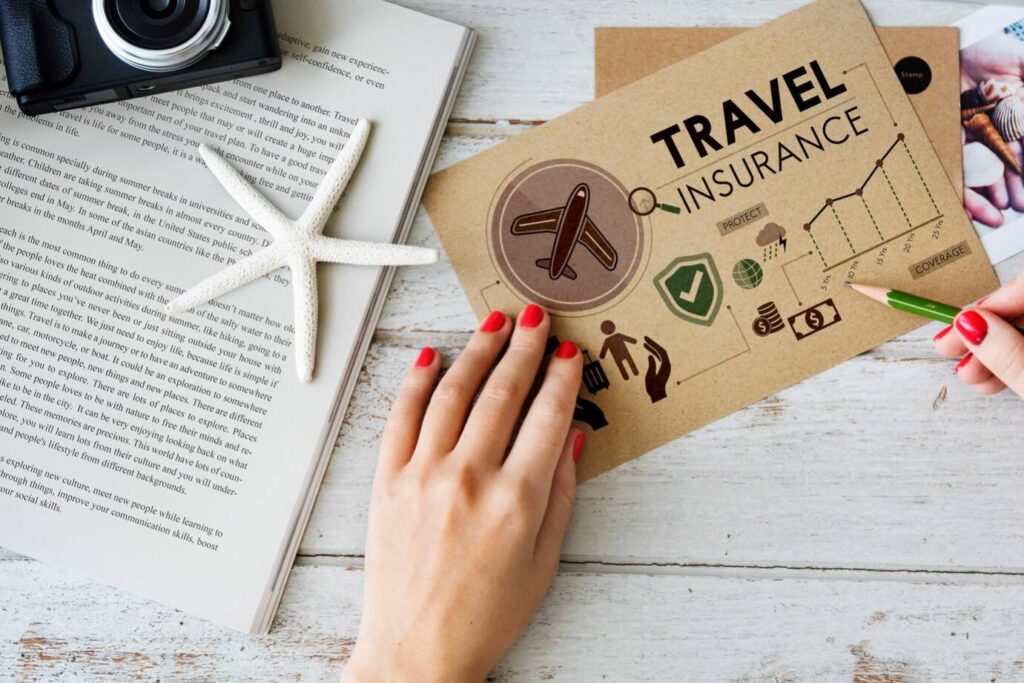 Person holding a pencil over a travel insurance document, with a camera, starfish, and open book on a wooden table.
