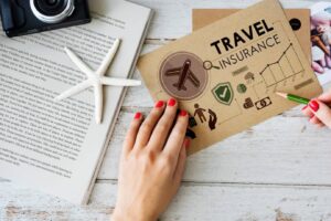 Person holding a pencil over a travel insurance document, with a camera, starfish, and open book on a wooden table.