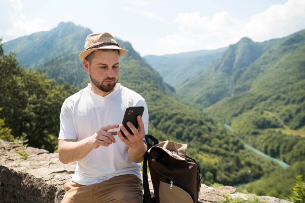 Man in a straw hat using a smartphone outdoors with a mountainous backdrop.