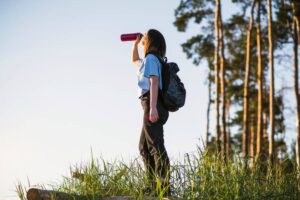 Young woman hiking, drinking water from a bottle, standing near trees under a clear sky.