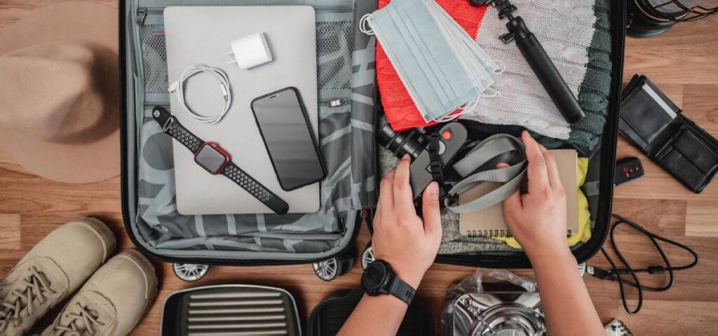Overhead view of a person packing a suitcase with clothes, electronic devices, and travel essentials, including a facemask.