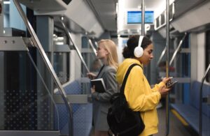 Young woman in a yellow jacket with headphones using a phone on a metro, with another commuter in the background.