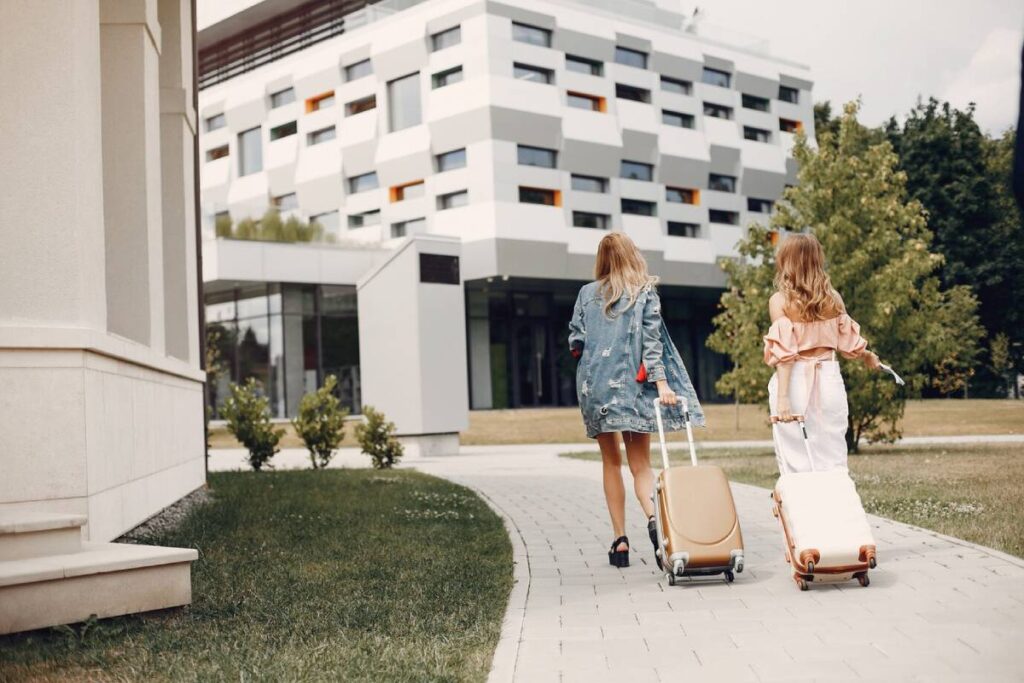 Two women walking with suitcases towards a modern building on a sunny day.