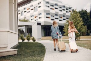 Two women walking with suitcases towards a modern building on a sunny day.