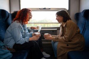 Two women joyfully chatting and holding coffee cups while sitting opposite each other on a train.