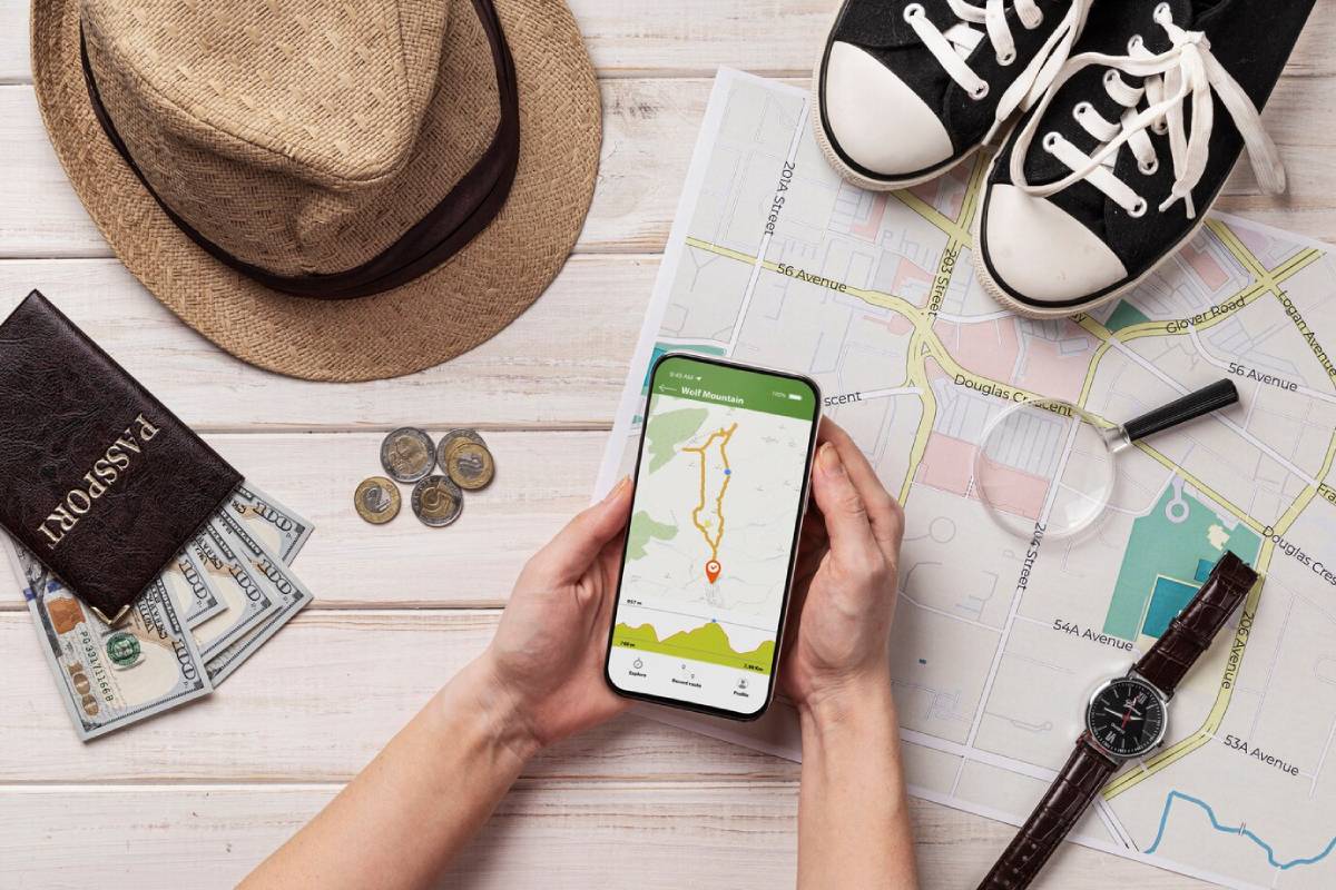 Hands holding a smartphone displaying a map, surrounded by travel essentials like a hat, shoes, passport, money, and a watch on a wooden surface. 