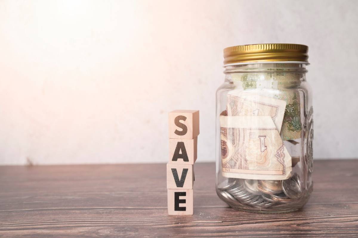  Glass jar filled with coins and banknotes next to wooden blocks spelling SAVE on a table.