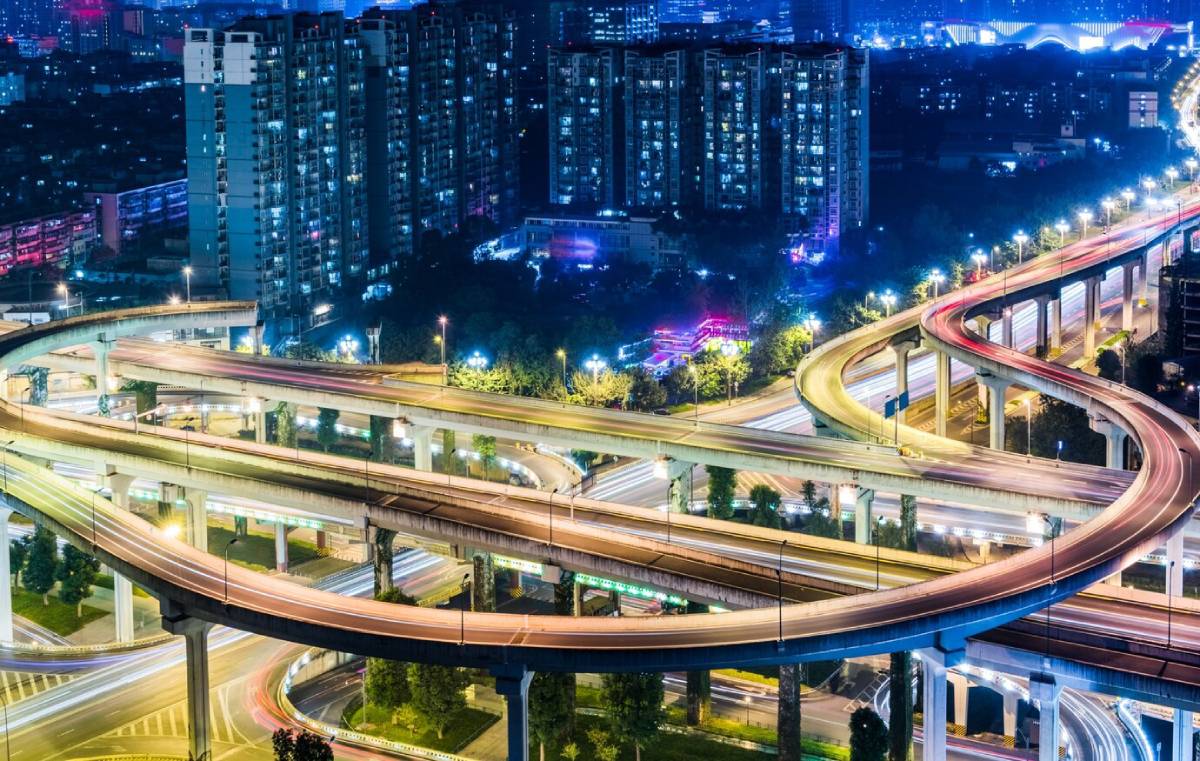 Aerial night view of a brightly lit, complex multi-level road interchange in a densely populated urban area.