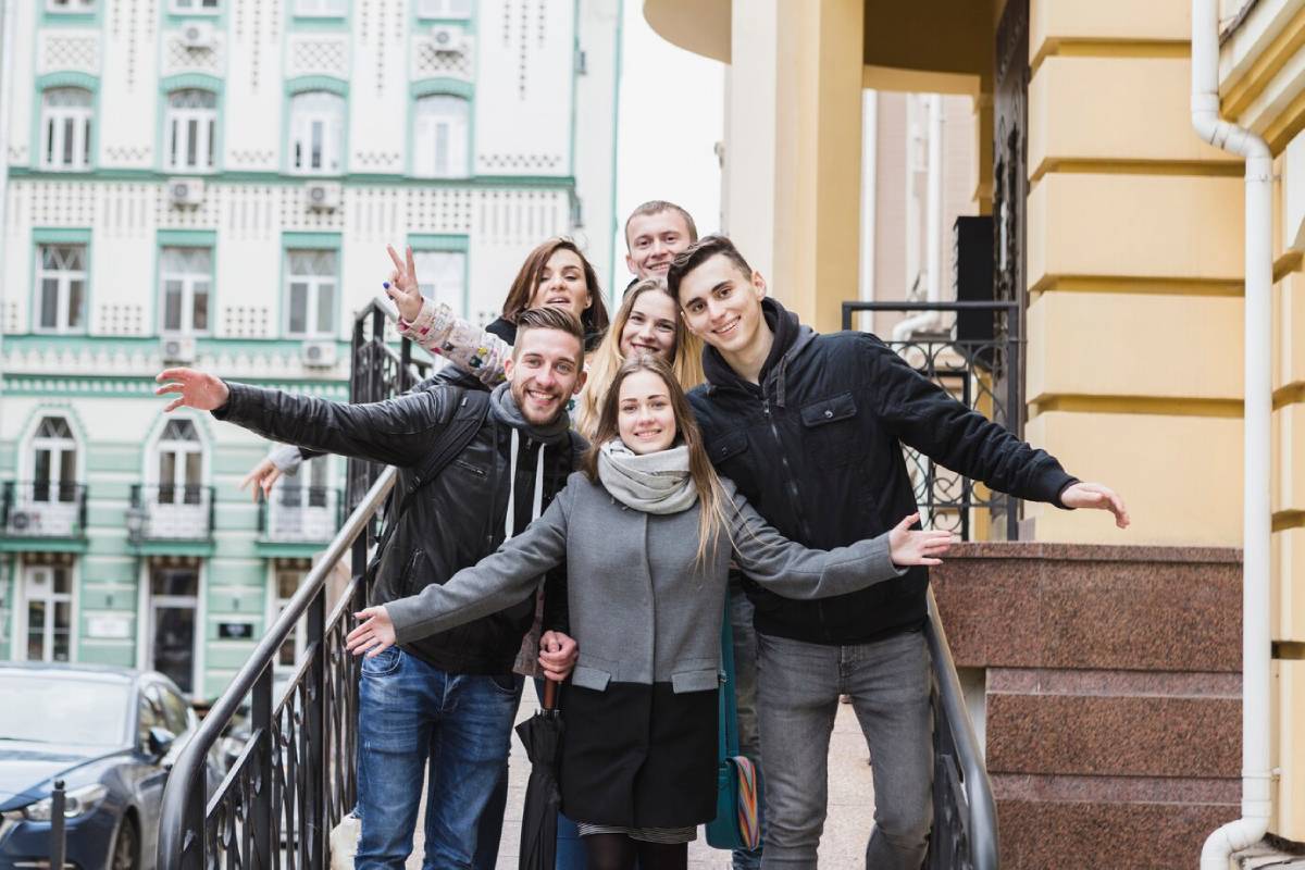 Group of young friends posing cheerfully on urban stairs, with ornate building in the background. 
