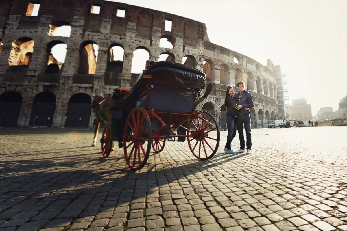 Couple stands near a horse-drawn carriage in front of the Colosseum in Rome at sunrise. 