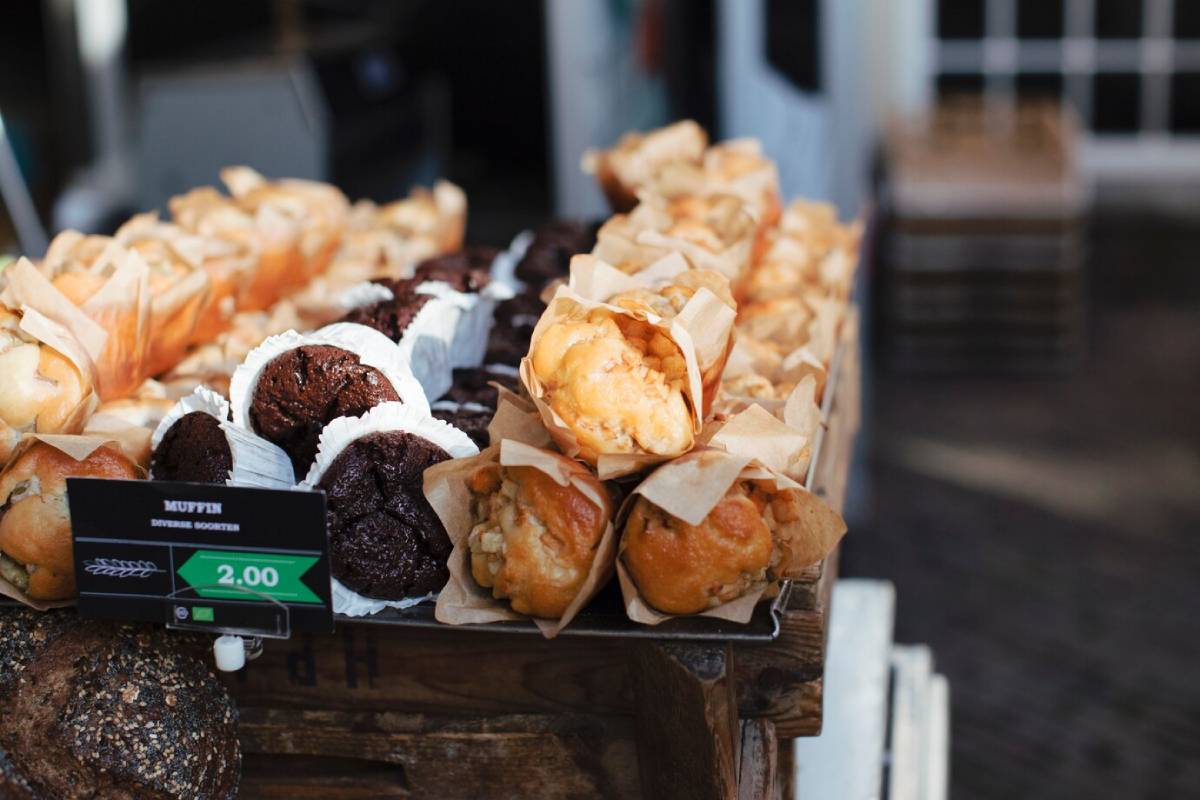 Assorted muffins displayed for sale at a market stall, with prices shown on a digital sign.
