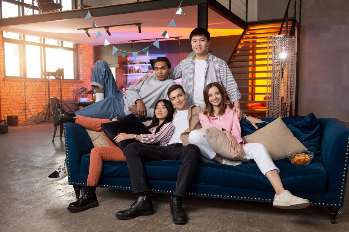 Five young adults smiling and sitting together on a blue sofa in a stylish loft with festive decorations. 