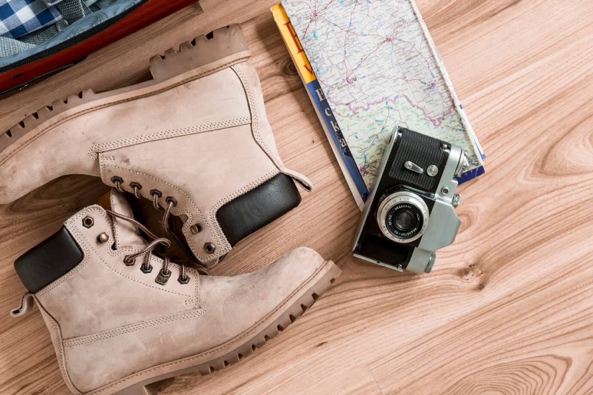 Pale hiking boots, a folded map, and a vintage camera on a wooden floor, suggesting travel planning. 