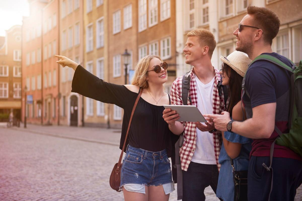 Four friends exploring a city street, one pointing out something of interest while another holds a tablet. 