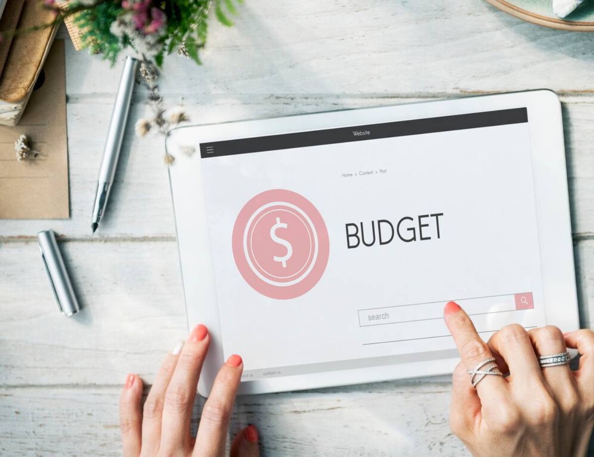 Hands using a tablet displaying a budget management website, with pens and decorations on a wooden table.