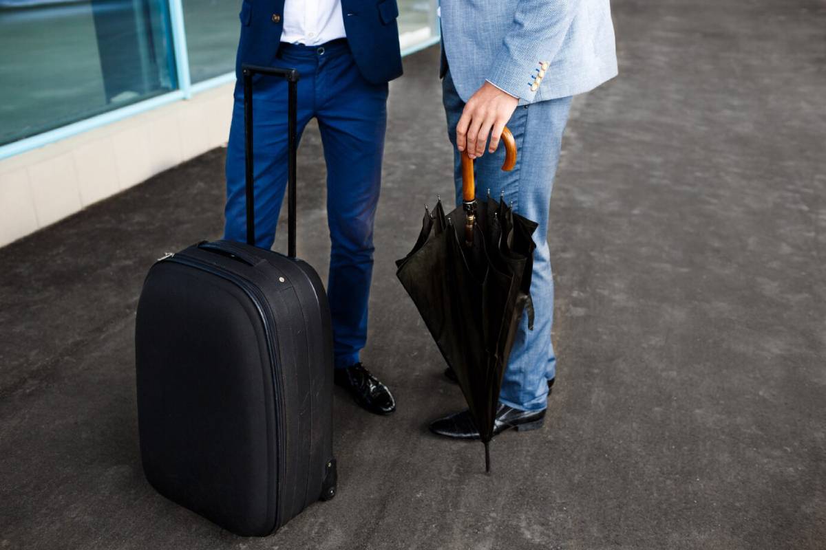 Two men in suits stand together, holding luggage and umbrellas, ready for travel in a rainy setting.
