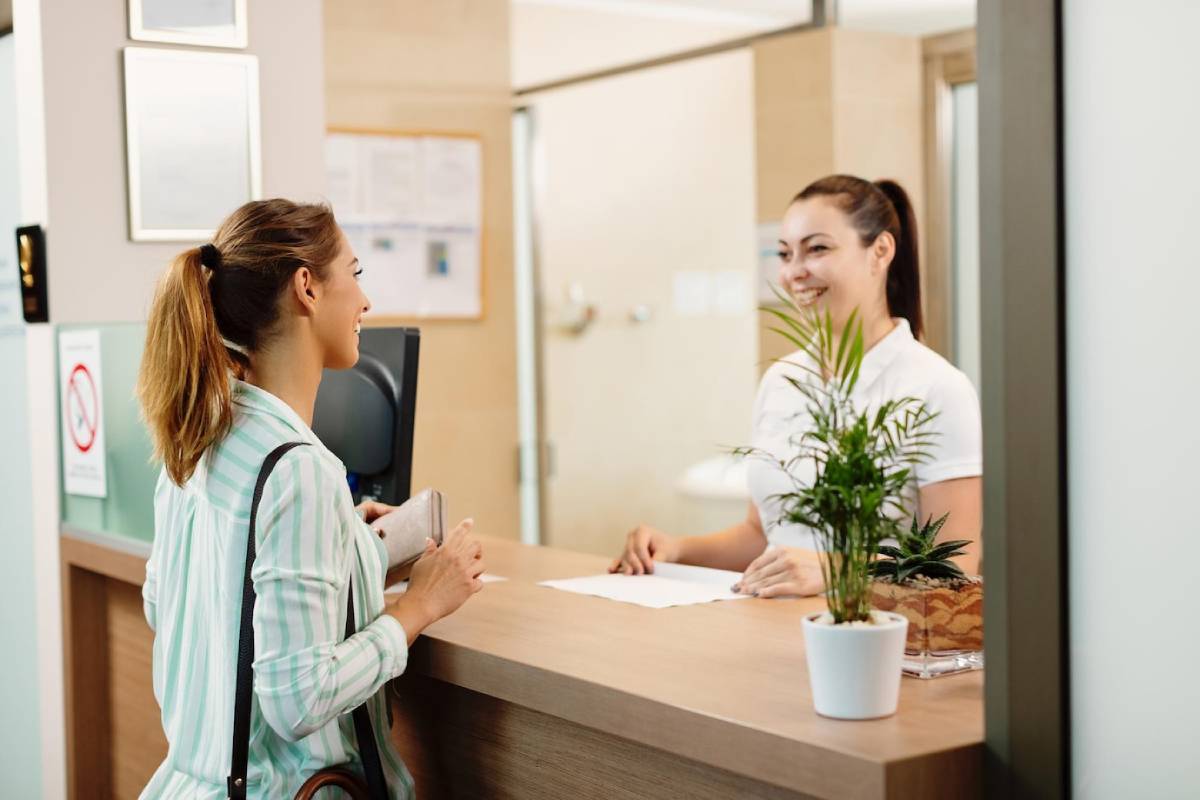  A woman checks in at a reception desk, smiling at the receptionist in a clinic.
