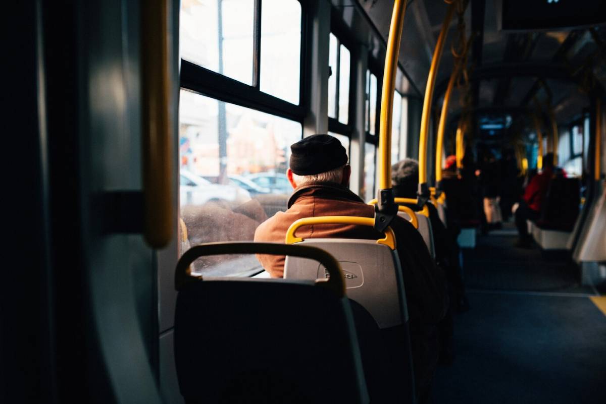 Passengers seated on a city bus during a bright day, viewed from behind. 