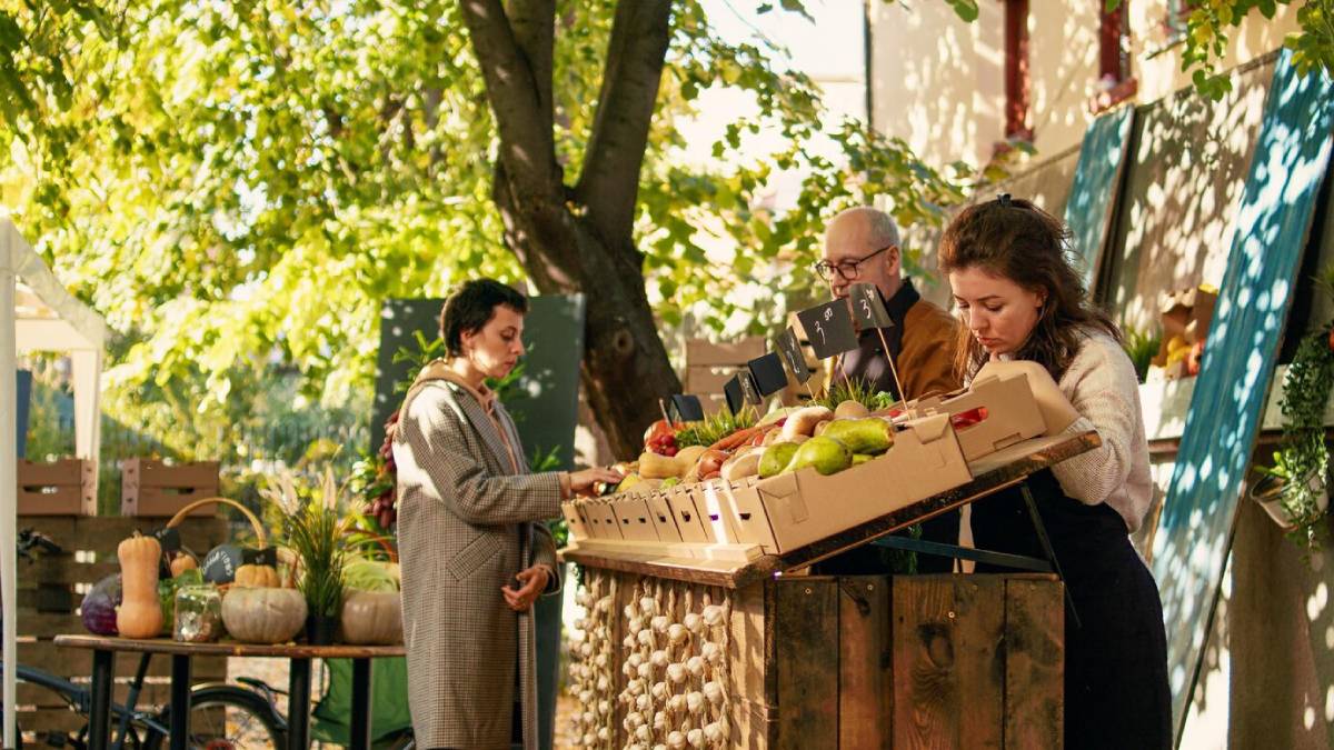 People shopping at a local outdoor market with fresh fruits and vegetables on display under a tree. 