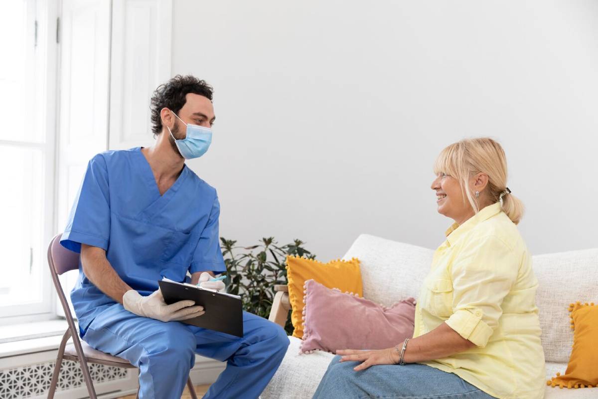 Male nurse wearing a mask consults with smiling elderly woman in a bright living room. 