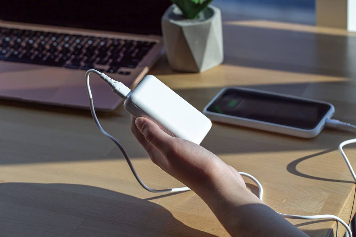  A person holding a portable charger connected to a smartphone and laptop on a wooden desk.