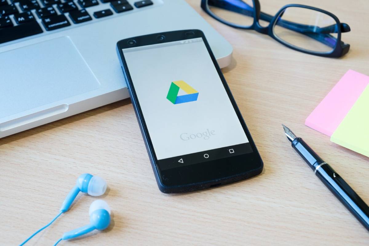  Smartphone with Google Drive logo on screen beside a laptop, glasses, and sticky notes on a wooden desk.