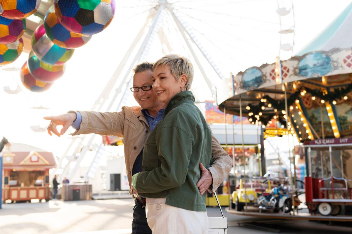  A happy older couple embracing at an amusement park, with a Ferris wheel and festive lights in the background.
