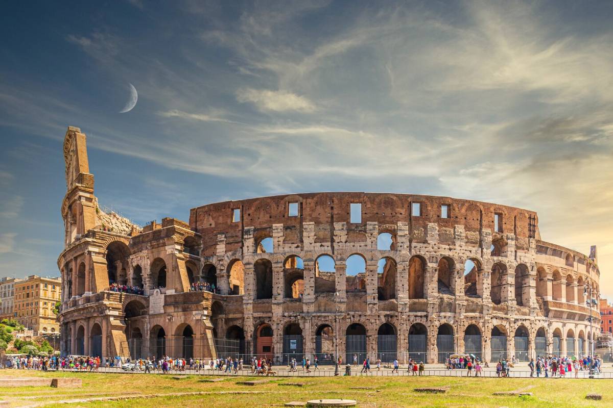 The Colosseum in Rome on a bright day, showing the iconic exterior with tourists and a crescent moon in the sky. 