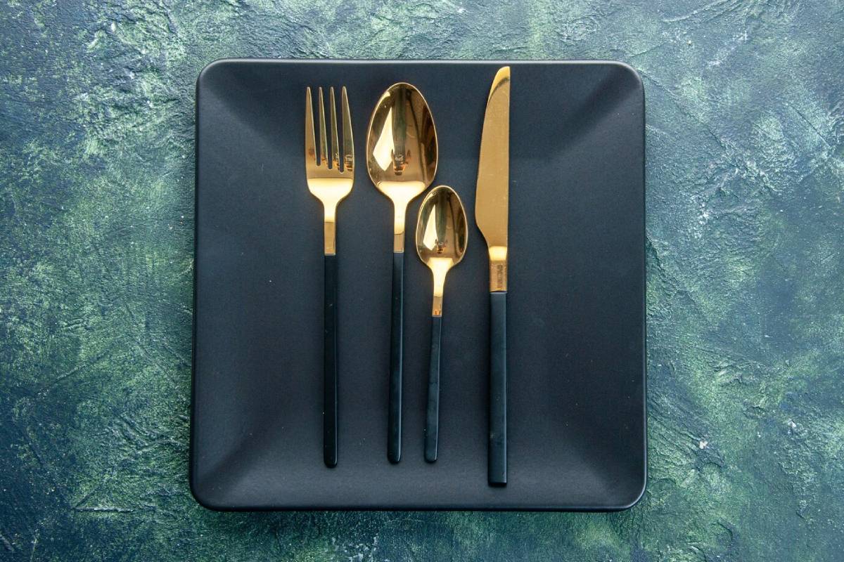 A black plate with a gold fork, knife, spoon, and teaspoon on a green background. 