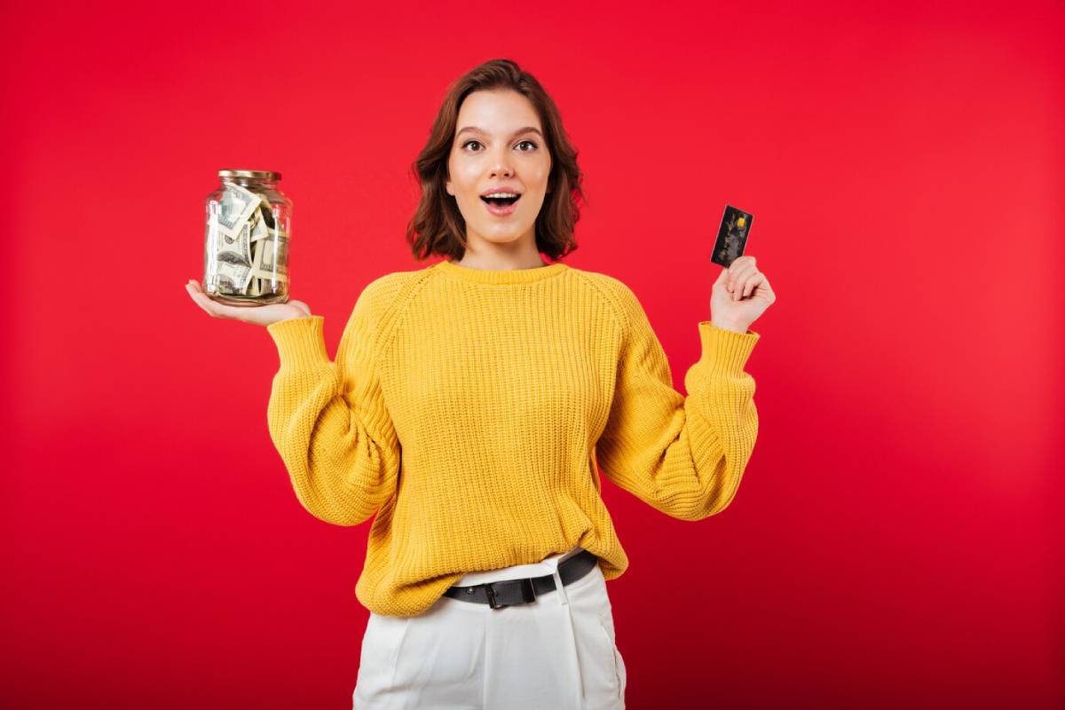  Woman in yellow sweater holding a jar of money and a credit card, with a surprised expression, against a red background.