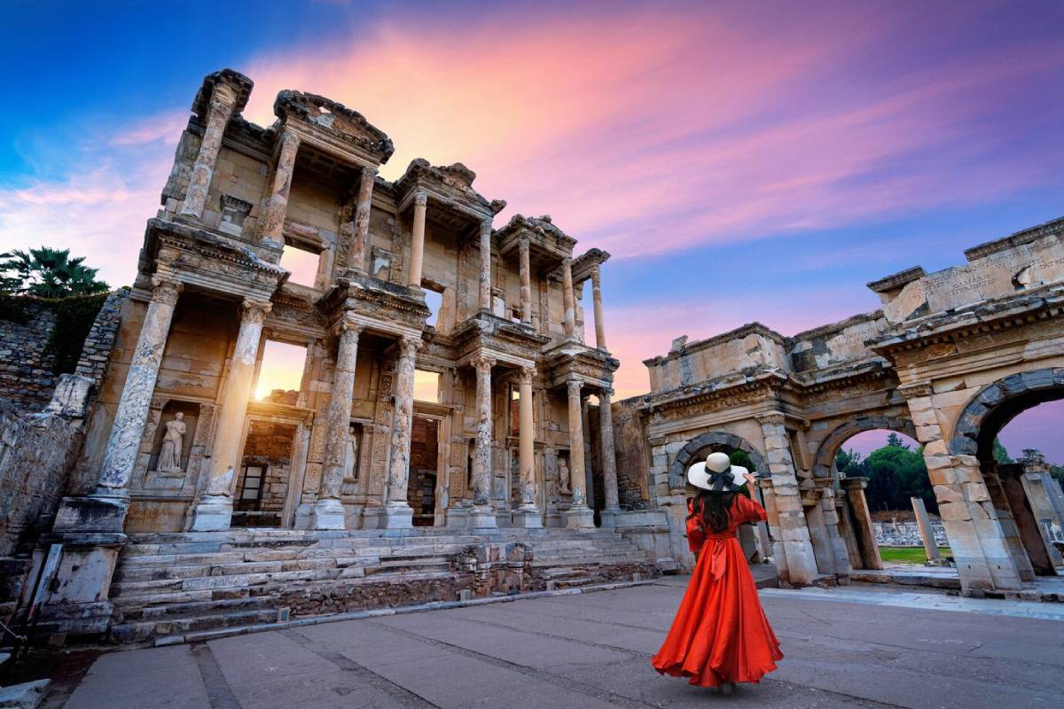 Woman in red dress and hat admires ancient ruins at sunset. 