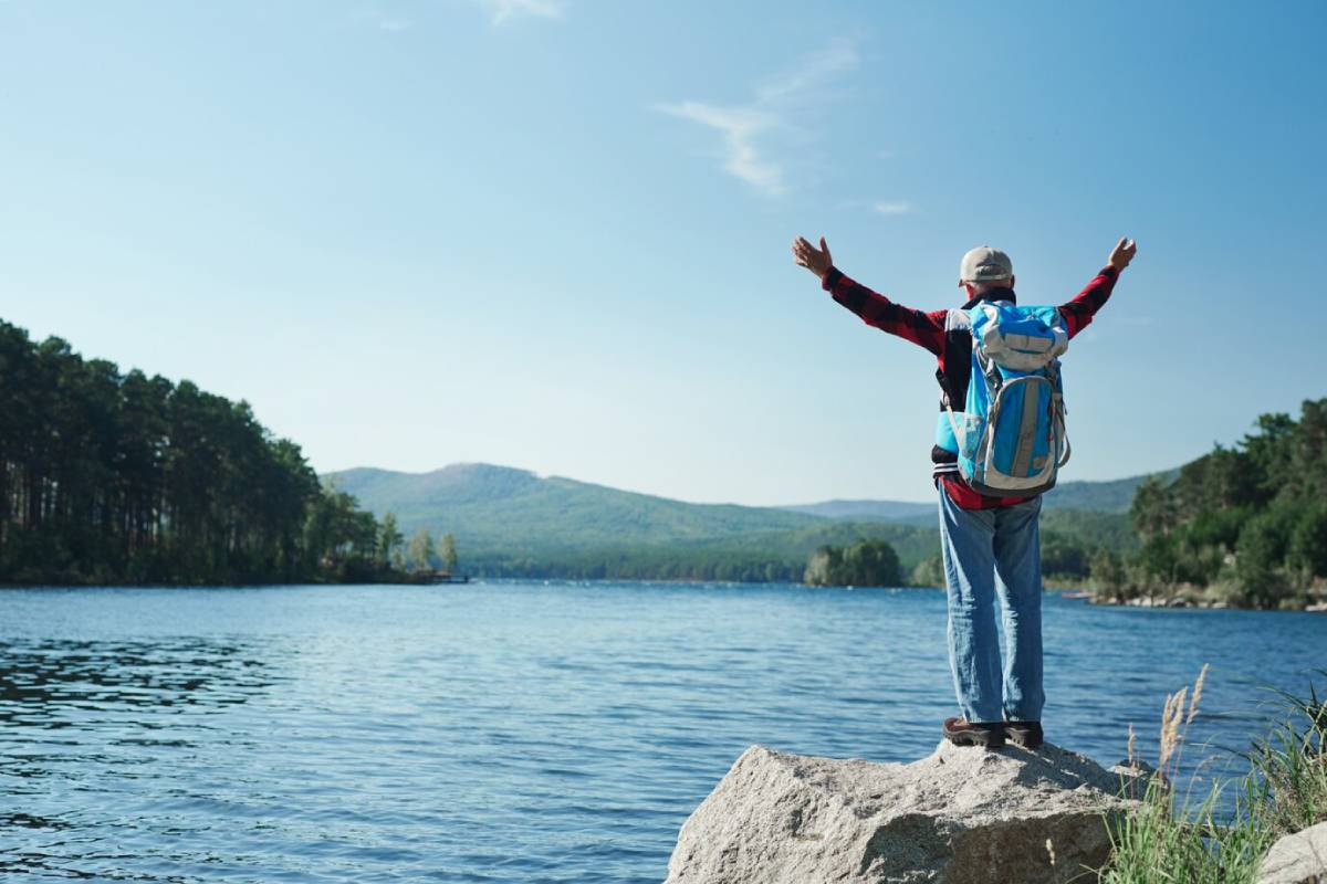 Person with backpack standing on a rock by a lake, arms raised in joy, with mountains in the background.