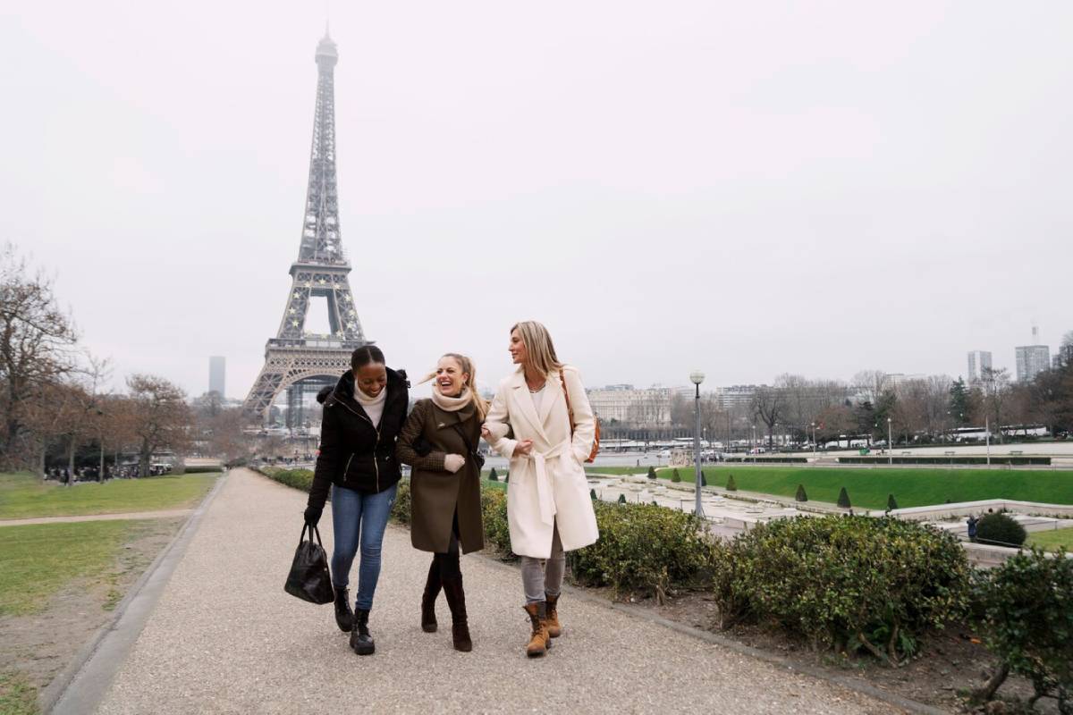 Three women walking and laughing near the Eiffel Tower on a cloudy day.