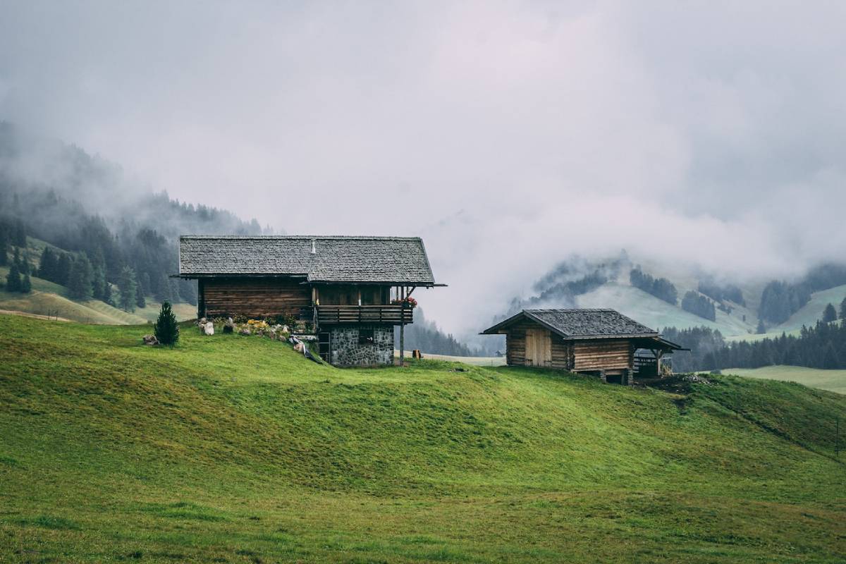  Two wooden cabins on a misty green hillside, with fog-blanketed mountains in the background.