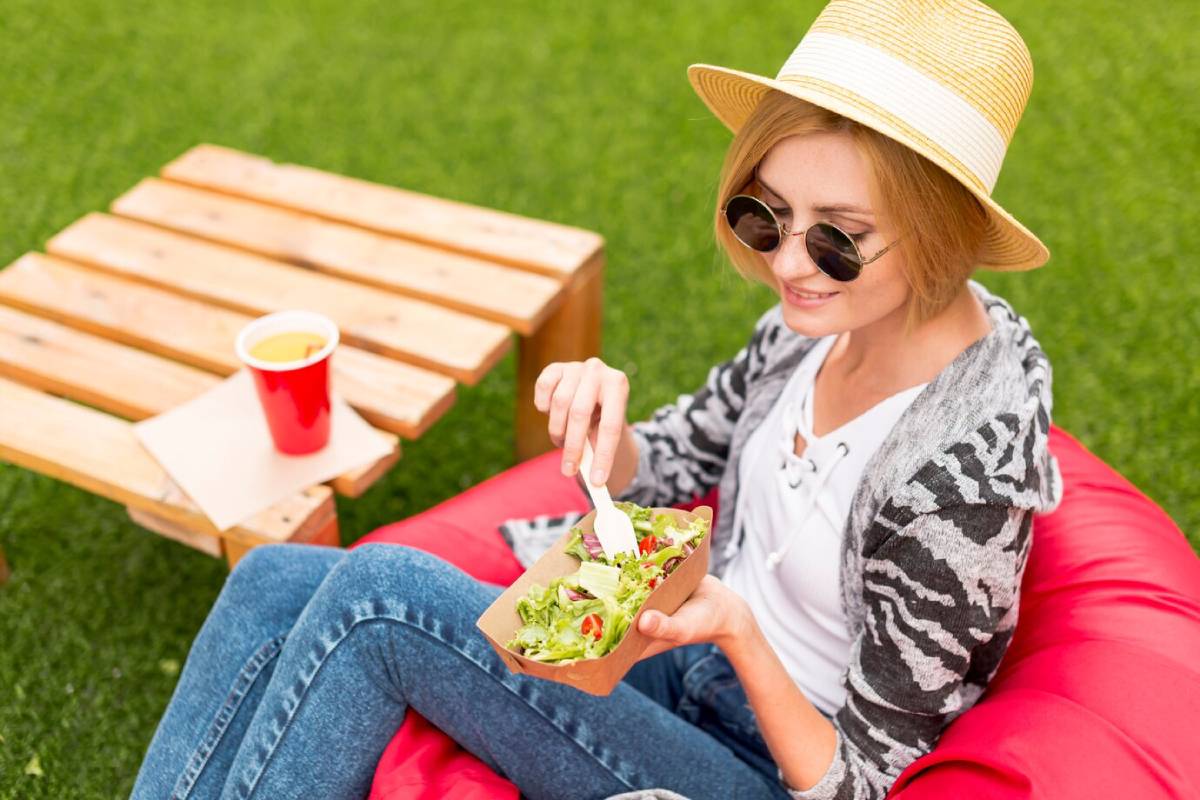 Woman in sunglasses and hat sitting on red bean bag outdoors, eating salad near a wooden table with red cup. 