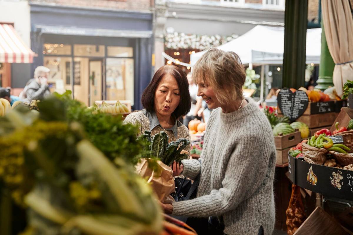 Two women examining vegetables at a busy indoor market, sharing a joyful moment.
