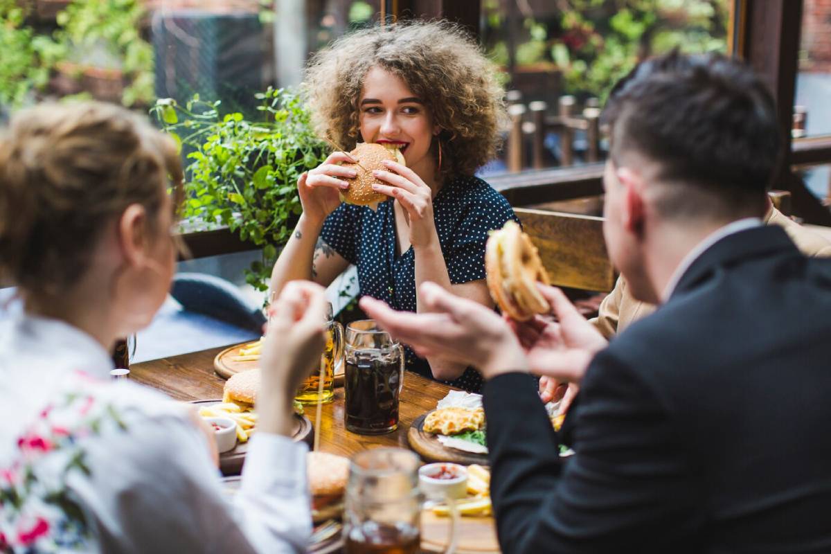  Three people enjoying burgers and drinks at an outdoor cafe table surrounded by greenery.