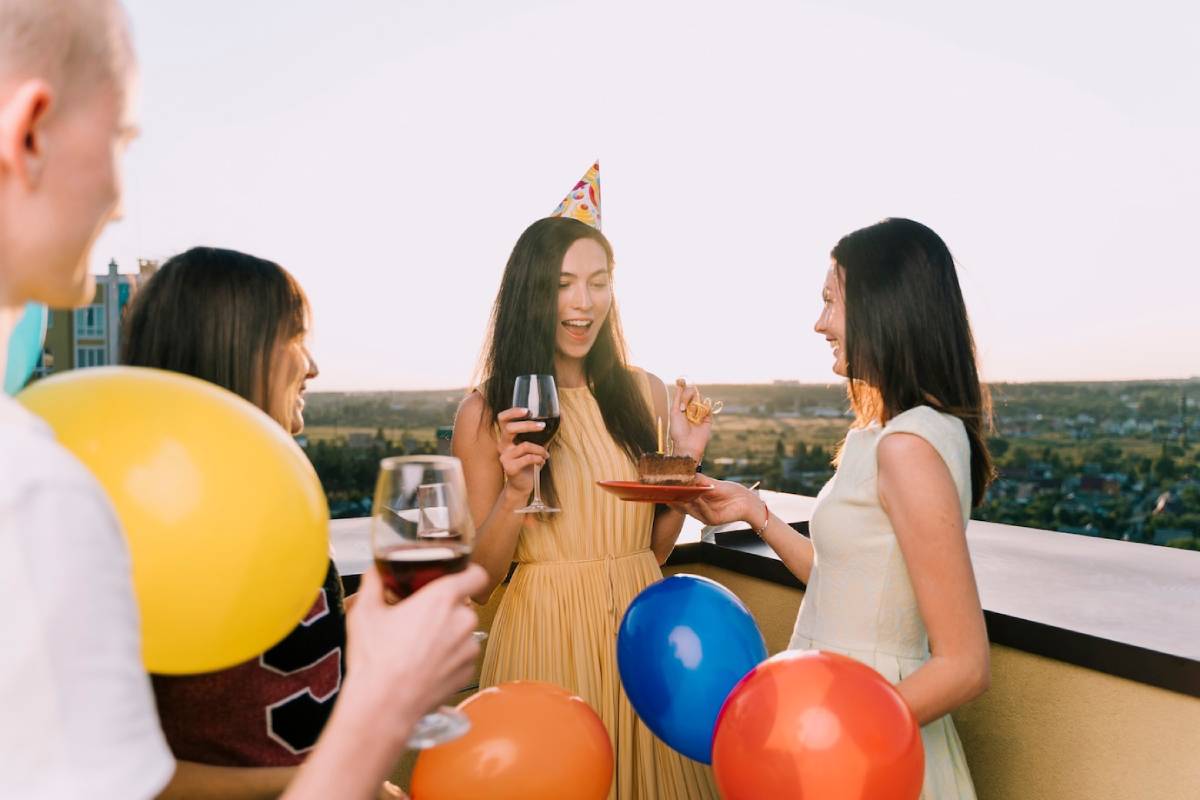 Four people celebrating with wine and cake at a rooftop birthday party, decorated with colourful balloons. 