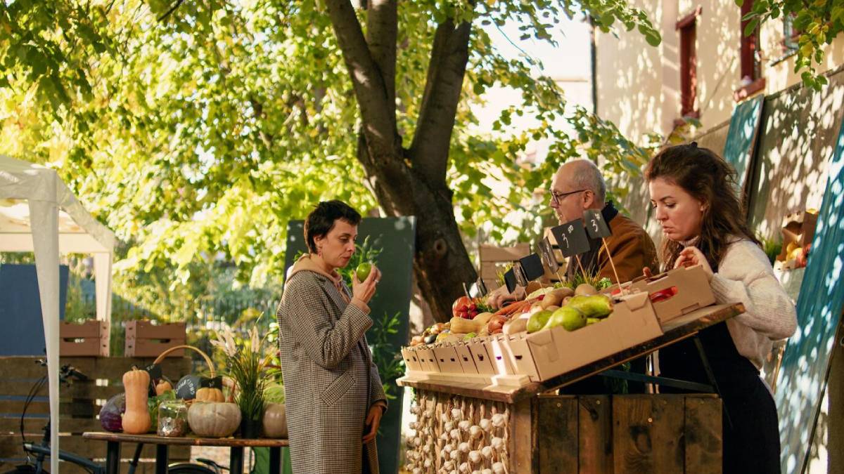 Three people shopping at a market stall under a tree. Fresh fruits and vegetables displayed on a sunny day. 